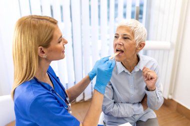 Doktor, kıdemli hastanın boğazını tahta dil basacağıyla muayene ediyor. Sağlık ve tıp konsepti. Mavi üniformalı ve eldivenli kadın hemşire sağlık kontrolü yapıyor. Boğaz ağrısı teşhisi.