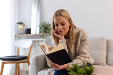 Koltukta kitap okuyan gülümseyen kadın, Mutlu Ev Rahatlığı ve Hobi Konsepti. Parlak, modern oturma odasında romanının tadını çıkarırken rahat süveter giyen çekici sarışın kadın..