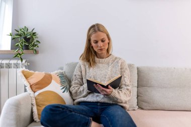 Kanepede kitap okuyan kadın, rahat ev rahatlığı konsepti, hafta sonu hobi yaşam tarzı. Sıcak örgü kazaklı çekici bir kadın koltukta minderle oturuyor, evde edebiyattan keyif alıyor..