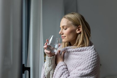 Hidrasyon ve Sağlık Konsepti. Evde su içen kadın, Sağlık ve Tıp konsepti. Sıcak battaniyeye sarılı çekici bir kadın bir bardak su tutuyor ve iyileşme sürecinde pencereden dışarı bakıyor..