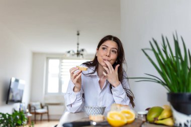 Lezzetli bir aperatifin tadını çıkaran ve ekmeğini yerken parmağını yalayan genç bir kadın. Beyaz gömlekli bir kadının güneşli bir evde sağlıklı bir kahvaltı yaptığı samimi bir yaşam tarzı..