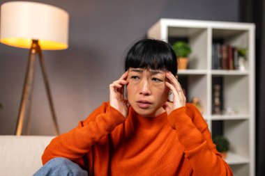 Başımda ağrı var. Migren ve baş ağrısı, sağlık sorunu olan insanlar, sağlık ve tıp konsepti. Kanepede oturan yorgun Asyalı kadın elleriyle acı dolu tapınakları tutuyor. Stres ve gerginlik.