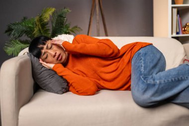 Hasta Asyalı Kadın Şiddetli Baş Ağrısı, Migren ve Yorgunluk Konsepti, Evde Sağlık Hizmetleri. Acı ve stresten muzdarip olgun bir kadın turuncu süveter içinde koltukta dinleniyor..