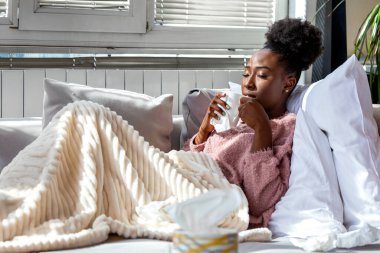 Soğuk için sıcak çay içiyorum. Kanepede yatan hasta Afrikalı Amerikalı kadın mevsimsel grip sorunu olan insanlar, Sağlık ve Tıp Konsepti. Battaniyeye sarılmış siyah bir kadın kupa ve doku tutuyor..