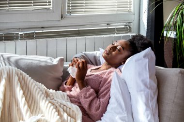 Griple uyuyorum. Kanepede dinlenen hasta Afrikalı Amerikalı kadın, solunum hastalığı olan insanlar, Sağlık ve Tıp konsepti. Yorgun, siyah bir kadın battaniyeye sarılmış, elinde doku tutuyor..