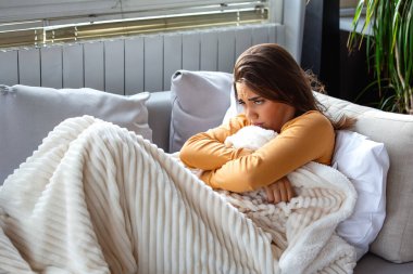 Ateşli hasta kadın kanepede titriyor. Beyaz battaniyeye sarılmış, üzgün bir ifadeyle yüzünü çeviren, üşümüş ve kötü hisseden hasta bir kadın. Grip ve mevsimsel virüs için sağlık ve tıp konsepti.