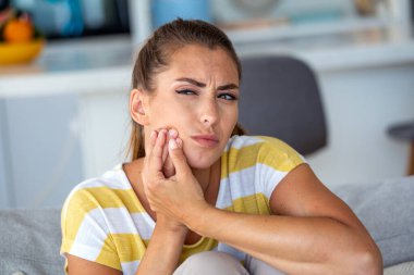 Hassas diş ağrısı, diş ağrısı olan genç kadın, diş bakımı sorunu olan insanlar, sağlık sigortası ve ilaç konsepti. Çekici kadın kanepede oturuyor ve acı veren çenesini eliyle tutuyor.