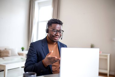 Bir dizüstü bilgisayardaki video görüşmesine katılan kulaklık takan Afro-Amerikalı bir adam. Profesyonel erkek jest yaparken konuşur, müşteri destek ajanı olarak çalışır ya da ev ofisinden serbest çalışır..
