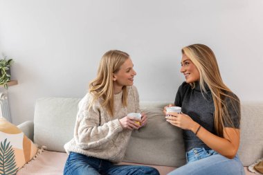 İki genç ve güzel kadın kanepede oturup kahve içerken sohbet ediyorlar. Mutlu arkadaşlar sıcak bir oturma odasında sohbet ediyorlar. Kapalı alanda arkadaşlık ve iletişim kavramı.