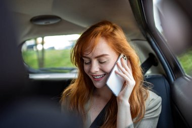 Arabanın arka koltuğunda akıllı telefondan konuşan mutlu, genç, kızıl bir kadın. Kızıl saçlı kız takside seyahat ederken cep telefonu kullanıyor ve gülümsüyor. İletişim ve teknoloji yaşam tarzı kavramı.