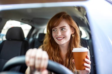 Elinde kahve fincanı olan dikkati dağılmış bir kadın sürücü yoldan uzağa bakıyor. Otomotiv sigortası mesuliyeti, tehlikeli sürüş alışkanlıkları ve modern bir arabada sabah trafiği riskleri..