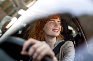 Araba kullanan kadın gülümsüyor. Direksiyondaki kızıl saçlı kadın sürücü. Otomotiv Sigortası, Güvenli Sürüş ve Seyahat Konsepti. Modern Araçtaki Genç Kadın.