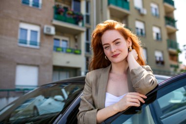 Modern bir şehirde, siyah bir arabanın yanında duran, kendine güvenen bir gülümsemesi olan, parlak güneşli bir günde şık bir ceket ve altın küpeler takan profesyonel, genç, kızıl saçlı bir iş kadını.