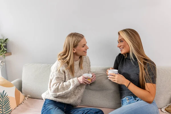 İki genç ve güzel kadın kanepede oturup kahve içerken sohbet ediyorlar. Mutlu arkadaşlar sıcak bir oturma odasında sohbet ediyorlar. Kapalı alanda arkadaşlık ve iletişim kavramı.