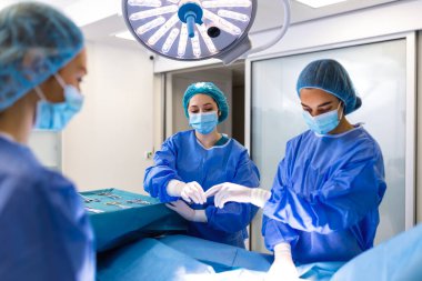 Mavi önlüklü ve maskeli kadın cerrah ve asistanlardan oluşan bir ekip, parlak ameliyathane ışıkları altında steril bir tepside cerrahi aletler hazırlıyor, klinikteki tıbbi bir işlemden önce koordine oluyorlar.