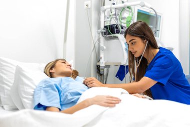 Mavi önlüklü genç bir kadın doktor ya da hemşire hastane yatağında yatan bir hastayı muayene etmek için steteskop kullanıyor. Profesyonel tıbbi konsültasyon, teşhis kontrolü ve özenli klinik bakım..