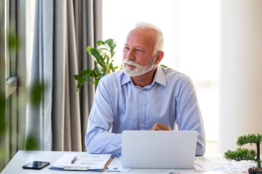 Üst düzey işadamı parlak ofiste dizüstü bilgisayarla çalışıyor. Profesyonel yaşlı adam düşünüyor, başka tarafa bakıyor, sağlık ve sağIık kavramı. Başarılı erkek girişimci bilgisayarla masada oturuyor..