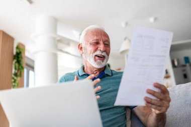 Kıdemli Adam Düşük Kamu Hizmetleri Tasarısı, Finansal Başarı ve Emeklilik Konsepti hakkında mutlu. Gri sakallı, elinde kağıt olan yaşlı bir erkek ve parlak odada neşeli bir ifadeyle laptop ekranına bakıyor..