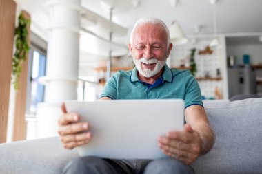 Son sınıf öğrencisi Tablet, İletişim ve Teknoloji Konseptine Görüntü Çağrısında Bulunacak. Evdeki kanepede otururken dijital cihaz ekranına bakıp gülen gri sakallı mutlu yaşlı bir erkek..
