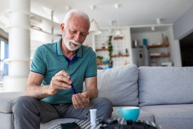 Diyabet Glikoz Testi. Kan şekeri seviyesi, sağlık sorunu olan insanlar, sağlık ve tıp konsepti. Yaşlı adam kanepede oturuyor ve evde test etmek için parmak üstüne kalem kullanıyor..