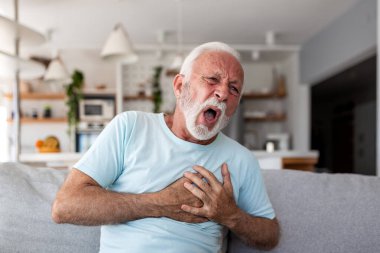 Kalp krizi ve göğüs ağrısı. Kardiyovasküler Hastalık, Kalp Sorunlu İnsanlar, Sağlık ve Tıp Konsepti. Evde acı dolu bir ifadeyle kanepede oturan yaşlı bir adam..