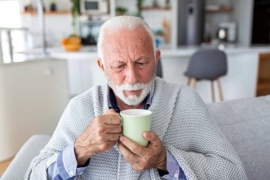 Son sınıf öğrencisi, battaniye, sıcak içecek, sağlık ve hastalık konseptiyle koltukta oturmaktan rahatsız oluyor. Parlak bir mutfakta gri örgü battaniyenin altında titrerken elinde bir kupa çay tutan yaşlı bir adam..