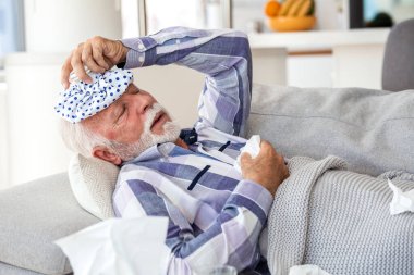 Migren ve yüksek ateş. Grip ve Soğuk, virüs enfeksiyonu olan insanlar, Sağlık ve Tıp konsepti. Yaşlı bir adam, gri bir battaniyenin altında titrerken elinde buz torbası tutarak kanepede yatıyor.