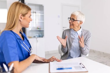 Profesyonel sağlık değerlendirmesi için klinikte yaşlı hasta sağlığıyla ilgili muayeneyi dinleyen mavi önlüklü kadın doktor.