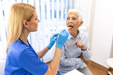 Kadın doktor ya da hemşire kıdemli bir hastanın boğazını kontrol ediyor. Sağlık ve tıp konsepti. Klinikte yaşlı bir kadının fiziksel muayenesi için dil bastırıcı kullanan profesyonel ya da doktor..