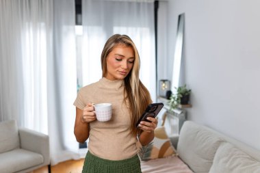 Oturma odasında elinde kahve fincanı tutarken akıllı telefon kullanan genç bir kadın. Modern Yaşam Tarzı ve Teknoloji Konsepti. Çekici Sarışın Kadın Evde Sosyal Medya Kontrol Ediyor. İletişim ve Gevşeme.
