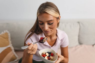 Sehpada Sağlıklı Kahvaltı 'da, Muz ve Böğürtlenli Yulaf Yiyen Genç Kadın' ın Yakın Çekimi. Besleyici meyve kasesinin, sağlığın ve beslenme konseptinin tadını çıkaran bir kadın, rahat bir oturma odasında oturuyor..