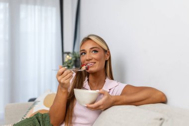 Kanepede Sağlıklı Kahvaltı Yapan Genç Kadın. Çekici Kadın Koltukta Oturuyor, Klozet Tutuyor ve Böğürtlenli Granola 'nın tadını çıkarıyor. Sabah Sağlığı, Beslenme ve Evde Yaşam Tarzı Konsepti.