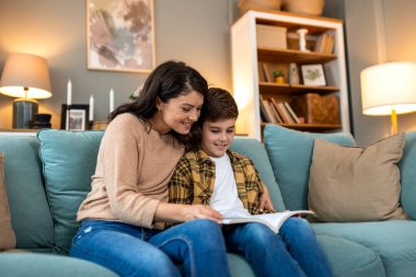 Anne ve oğlu kanepede kitap okuyor. Eğitim, hikaye anlatma ve aile bağları kavramı. Genç bir çocukla oturan mutlu kadın evdeki rahat oturma odasında kitap okuyor, birlikte güzel vakit geçiriyorlar..