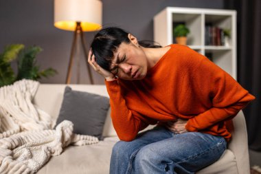 Kanepede karın ağrısı ve baş ağrısı çeken Asyalı bir kadın. Sağlık ve tıp konsepti. Karın ağrısı, mide bulantısı ya da regl sancısı çeken bir Japon kendini evde iyi hissetmiyor..