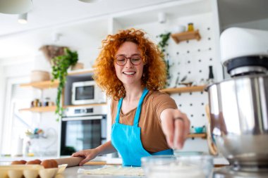 Mutfakta Kadın Pişirme, Yuvarlanan Hamur ve Hazırlama Gıda, Hobi ve Yaşam Tarzı Konsepti. Kıvırcık saçlı, önlük takan, hamura un tozu süren güzel bir kızıl kadın mikser ve tezgahın üzerinde yumurtalar..
