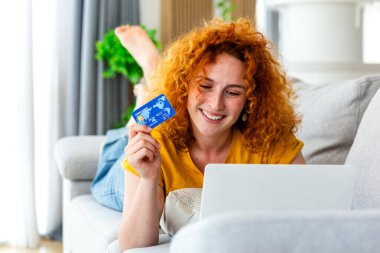 Kredi Kartıyla Mutlu Kadın Alışveriş İnterneti ve Kanepede Laptop. Genç Kızıl Saçlı Kız Ev 'de E-Ticaret Satın Alım yapıyor. Bankacılık, Finans, Tüketim ve Dijital Ödeme Teknolojisi.