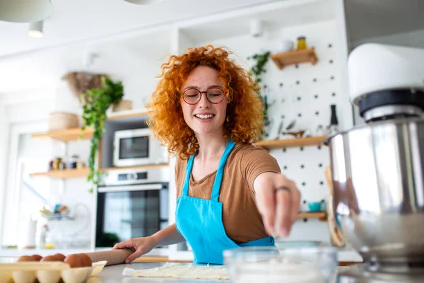 Mutfakta Kadın Pişirme, Yuvarlanan Hamur ve Hazırlama Gıda, Hobi ve Yaşam Tarzı Konsepti. Kıvırcık saçlı, önlük takan, hamura un tozu süren güzel bir kızıl kadın mikser ve tezgahın üzerinde yumurtalar..