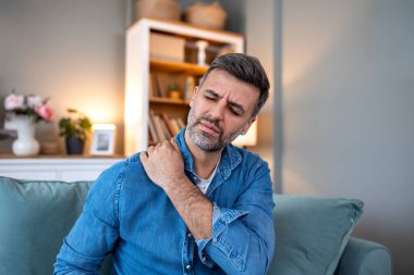 Omzum ağrıyor. Üst kol ağrısı, kas problemi, sağlık sigortası ve ilaç konsepti olan insanlar. Kanepede oturan ve elinle acı çeken bir omuz tutan adam. Kas sertliğinden muzdarip.