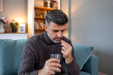 Evdeki kanepede oturup bir bardak suyla hap alan bir adam. Sağlık ve tıp konsepti. Grip ya da soğuk algınlığı belirtileri gösteren kişiler ilaç, vitamin ya da iyileşme için takviye maddeleri kullanmaktadır..