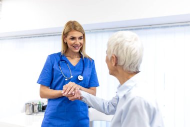 Klinikte yaşlı bir kadının elini tutan ilgili kadın doktor. Sağlık ve Tıp konsepti. Ofisteki tıbbi danışmanlık sırasında yaşlı hastayı destekleyen ve rahatlatan profesyonel hemşire..