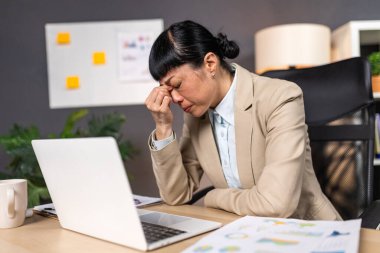 İş yerinde baş ağrısı çeken Asyalı yorgun bir iş kadını. Ofis masasında dizüstü bilgisayarı, gözleri kapalı, alnına dokunan bej ceketli stresli kadın çalışanı. Zihinsel sağlık tükenme kavramı.