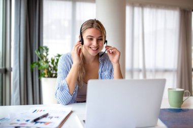 Bilgisayarda video görüşmesi sırasında kulaklık takan gülümseyen genç bir kadın, müşteri desteği ve uzaktan çalışma konsepti. Müşterilerle çevrimiçi konferans aracılığıyla iletişim kuran mutlu kadın serbest yazar ya da operatör.