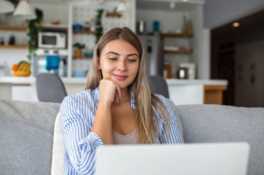 Evdeki koltukta dizüstü bilgisayar kullanan gülümseyen genç bir kadın, uzaktan çalışma ve yaşam tarzı kavramı. Oturma odasında oturan mutlu kadın serbest yazar mutfak geçmişi olan bilgisayar ekranına bakıyor..