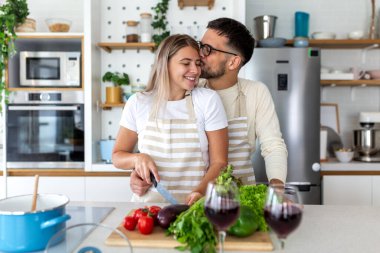 Modern Mutfakta Romantik Çiftler Birlikte Yemek Yapıyor. Aşk ve İlişki, Yiyecek ve İçki konsepti. Erkek, sağlıklı sebze yemeği hazırlarken kadını öpüyor. Önlükler içinde mutlu genç insanlar.
