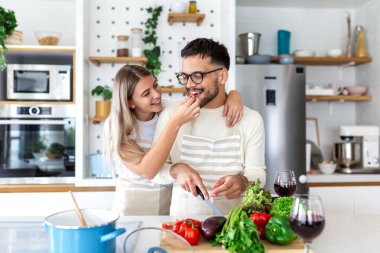 Yemek pişirirken Genç Mutlu Genç Çiftler Yemeği Tadına Bakıyor. Mutfakta eğlence, yiyecek ve içecek konsepti. Çekici kadın erkeği bir parça sebzeyle besliyor. İnsanlar evde birlikte sağlıklı yemek hazırlıyorlar..