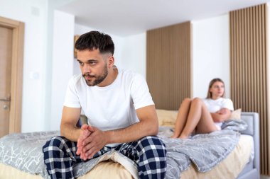 Üzgün adam tartışmadan sonra yatakta oturuyor. İlişki krizi konsepti. Arka planda bulanık bir kadın ve pijamalı mutsuz genç bir adam kendini yalnız hissediyor. İç mekanda anlaşmazlık var. Ruh sağlığı sorunları.