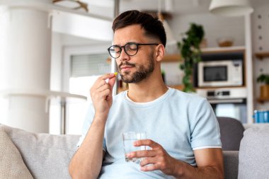 Genç adam, evde bir bardak suyla ilaç alıyor. Sağlık ve Tedavi Konsepti, Vitamin Emici veya Hastalık, Sağlık ve Sağlık Yaşam Tarzı İlacı.