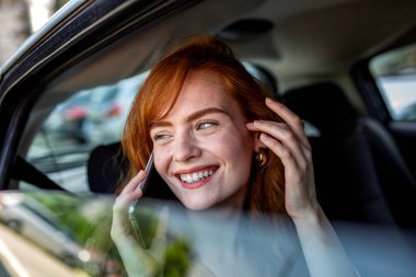 Arabada akıllı telefondan konuşan mutlu kızıl kadın. Genç ve çekici bir kadın seyahat ederken cep telefonu kullanıyor. İletişim ve yaşam tarzı konsepti. Kızıl kız telefonda gülümsüyor takside..