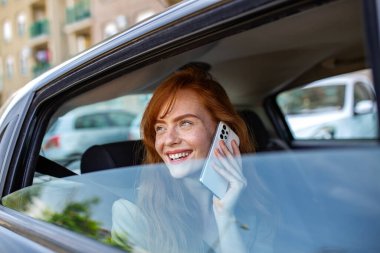 Kızıl saçlı kadın arabada telefonla konuşuyor. Çilli, akıllı telefon kullanan mutlu bayan yolcu. İletişim ve ulaşım konsepti. Güzel kızıl kız taksinin arka koltuğunda gülümsüyor..