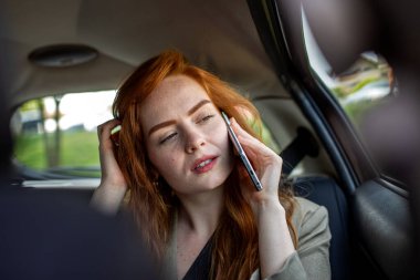 Kızıl saçlı kadın arabanın arka koltuğunda akıllı telefondan konuşuyor. Taksi sürerken cep telefonu kullanan kızıl saçlı profesyonel bir kadın. İş seyahati, iletişim ve modern yaşam tarzı kavramı.